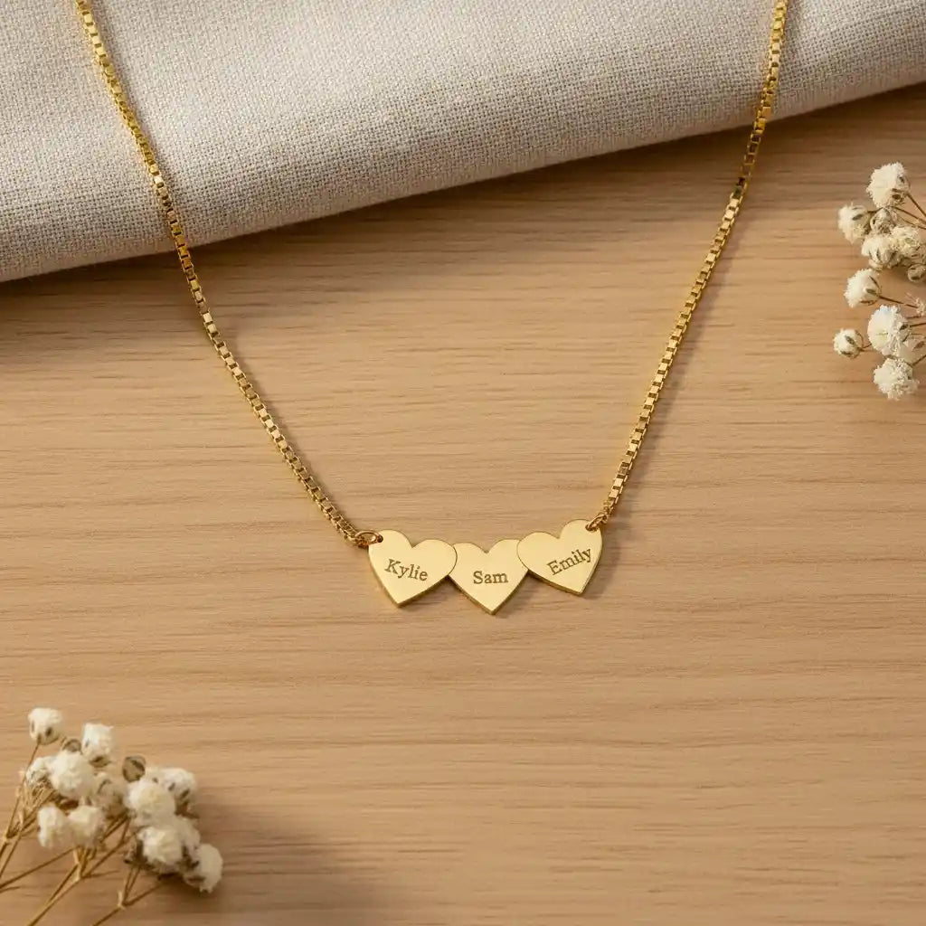 A gold chain necklace with three small engraved heart pendants reading 'Kylie', 'Sam', and 'Emily', styled on a light wood surface with a beige fabric napkin and small white flowers.