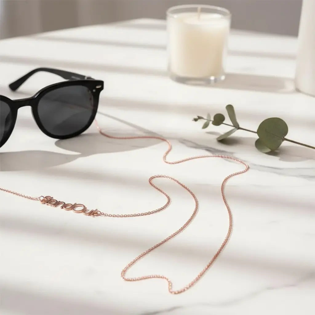 A rose gold chain necklace with the name 'SienaD'aki' spelled out in cursive script, styled on a white marble surface next to black sunglasses, a green eucalyptus sprig, and a lit candle.