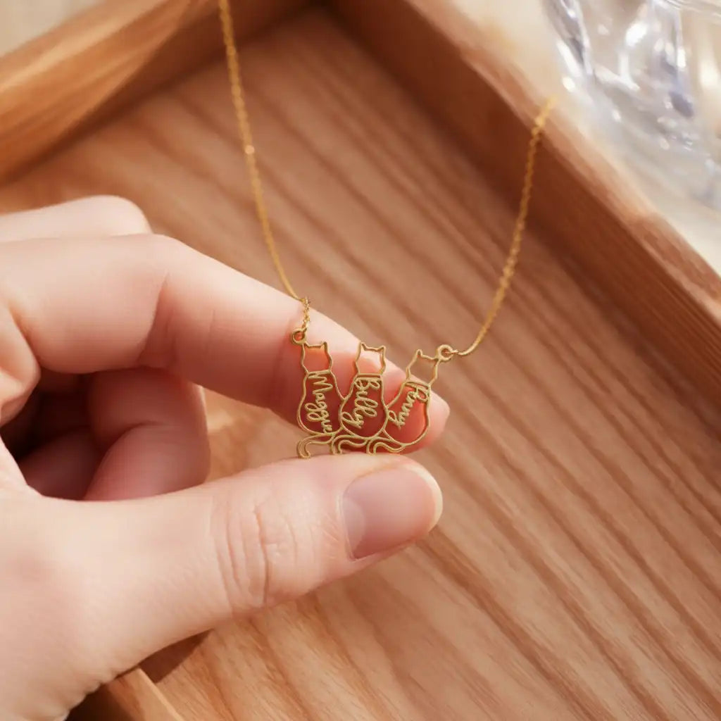 A close-up of a hand holding a gold necklace with three interlocking cat pendants, each engraved with a different script name, set against a warm wooden tray background.