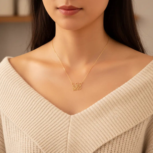 A close-up lifestyle shot of a woman wearing a delicate gold necklace with a script pendant spelling 'HBD' against a soft beige knit sweater.