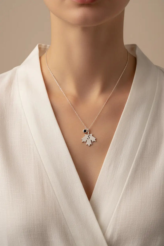 A close-up of a model wearing a delicate silver pendant necklace with a small black stone and a white leaf-shaped charm, styled with a white wrap dress against a soft beige background.