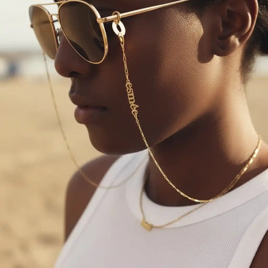 A close-up side profile of a person wearing round gold-framed sunglasses with a delicate gold chain strap hanging from the temple, along with layered gold necklaces and a white sleeveless top, set against a softly blurred sandy beach background.