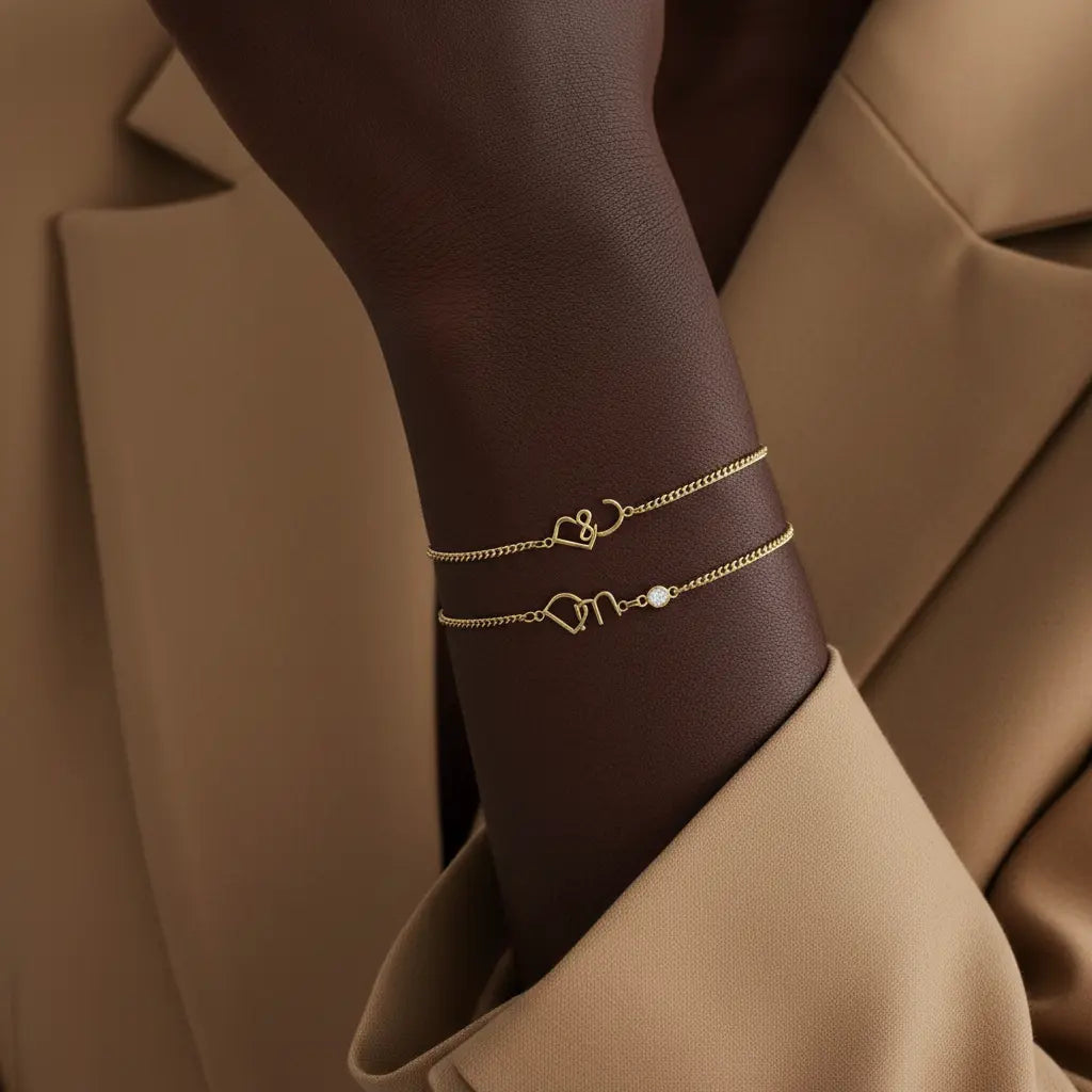Two delicate gold chain bracelets with custom script lettering and a small pearl accent, shown on a model's wrist against a beige blazer background.