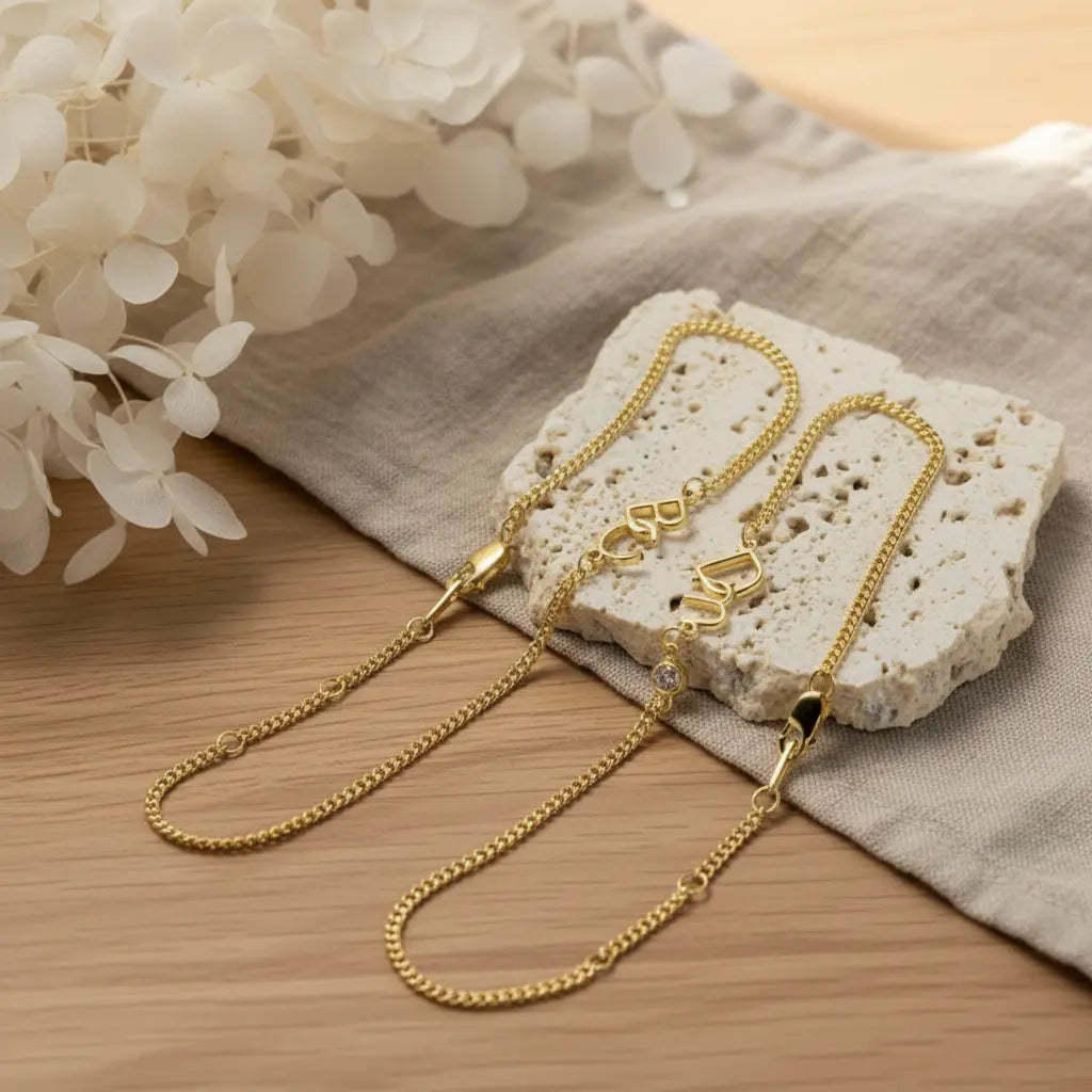 Two gold-tone chain necklaces with heart-shaped charms resting on a textured stone slab over a beige linen cloth and light wood surface, with white flowers in the background.
