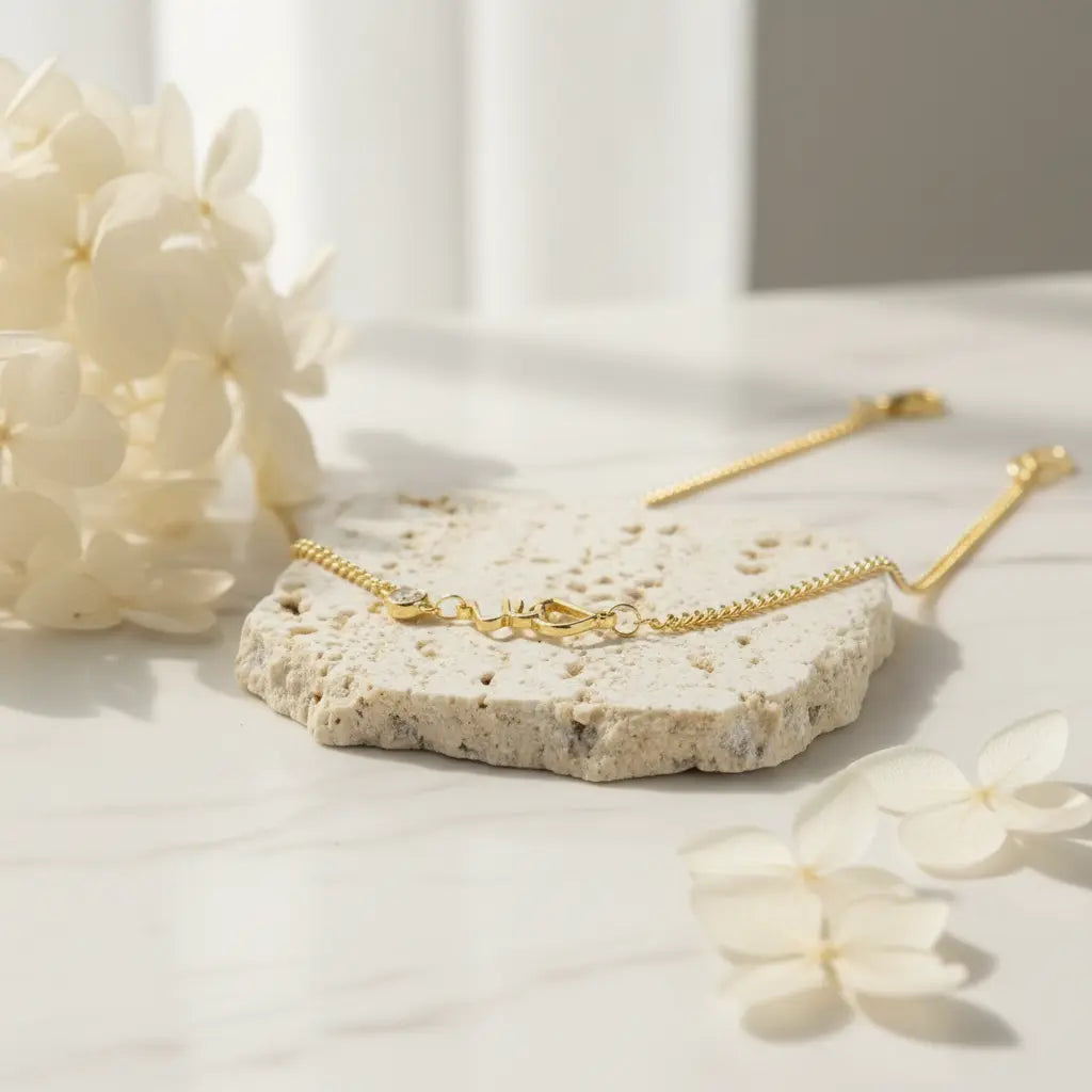 A delicate gold chain bracelet with a lobster clasp, displayed on a textured stone tray with white flowers on a light marble surface.