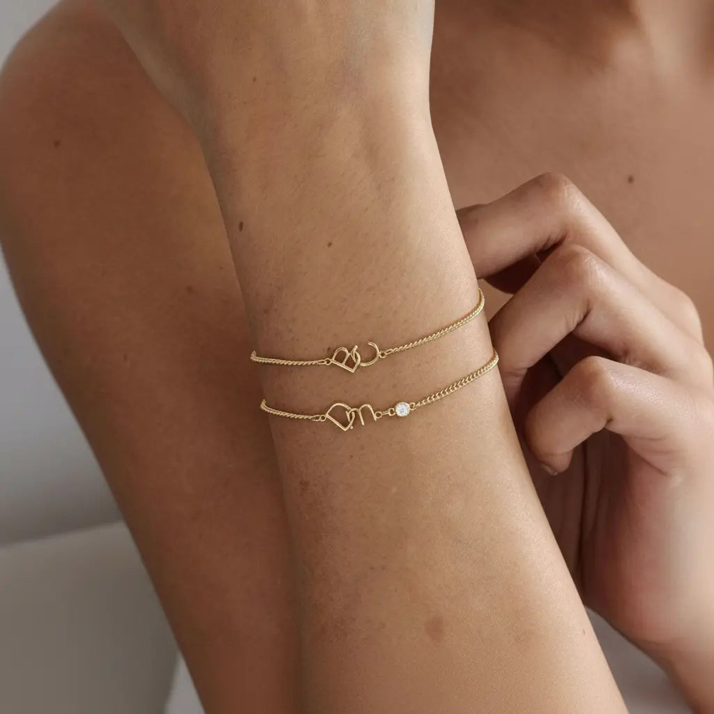 Two delicate gold chain bracelets with heart charms are shown worn on a person's wrist, with one bracelet featuring a small clear stone accent.